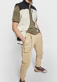 Man in beige cargobroek met ketting, olijfgroene t-shirt, witte en zwarte mouwloze vest, en multikleuren sneakers staat met handen in de zakken.