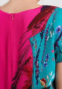 Bunte Bluse mit pinkem Rücken, türkisfarbenem Vorderteil mit Blumenmuster und pinkem Reißverschlussverschluss am hinteren Hals.