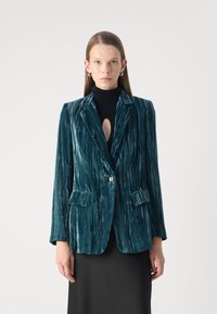 Teal velvet blazer with vertical ribbed texture, notched lapels, two front pockets, and a single button closure, paired with a black turtleneck.