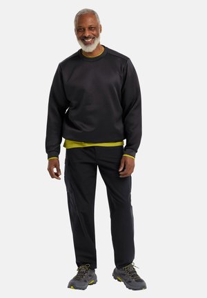 Felpa nera con un layer verde sottostante, abbinata a pantaloni cargo scuri e scarpe da trekking grigie con dettagli gialli.