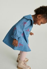 Denimkjole i blå farve, dekoreret med pink broderede sløjfer, krave og elastiske manchetter. Bæres med mønstrede pink strømper og metalguldsko.