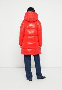 Rote Daunenjacke mit glänzender Oberfläche, ausgestattet mit einer großen Kapuze und horizontaler Steppung. Die Jacke reicht bis zur Mitte des Oberschenkels und ist mit blauen Jeans kombiniert.