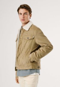 Corduroy jas in beige met een crèmekleurige schapenwol kraag. Bevat voorzakken, zichtbare naden en een getailleerde pasvorm. Gelaagd over een geruit shirt.
