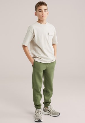 Zēns stāv ar rokām kabatās, valkājot bēša krāsas īss piedurkņu kreklu, olīvu zaļas jogger bikses un pelēkus sporta apavus uz neitrāla fona.