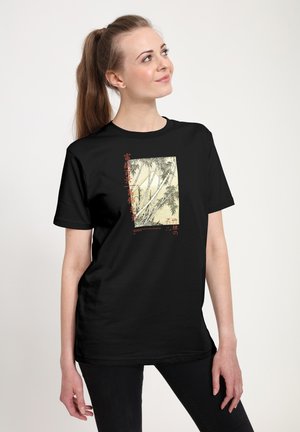 Junge Frau in schwarzem T-Shirt mit Bambus- und roter asiatischer Schriftkunst, steht und blickt leicht nach oben zur Seite vor weißem Hintergrund.