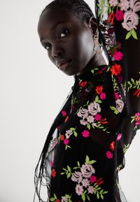 Top preto transparente com bordados florais vibrantes em rosa, vermelho e verde. Apresenta um decote alto e mangas longas e fluidas.