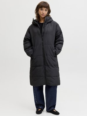 Donna che indossa un lungo piumino nero con cappuccio, mani nelle tasche, jeans a gamba larga blu scuro con pois bianchi e scarpe nere basse.