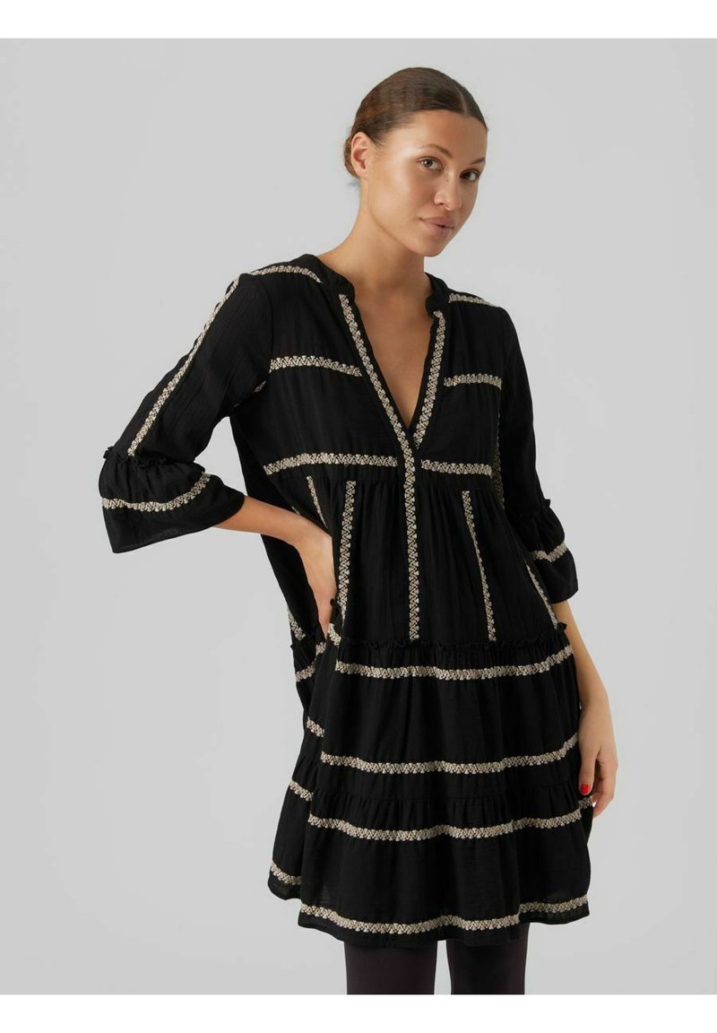 Robe noire avec un col en V et des manches trois-quarts. Présente des volants horizontaux et des détails de broderie décorative de couleur claire.