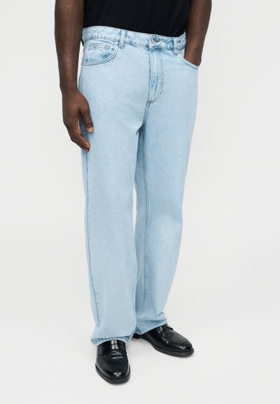 Mężczyzna w jasnoniebieskich jeansach o prostych nogawkach, z rękami w kieszeniach i czarnych mokasynach, stoi na tle jednolitej ściany.