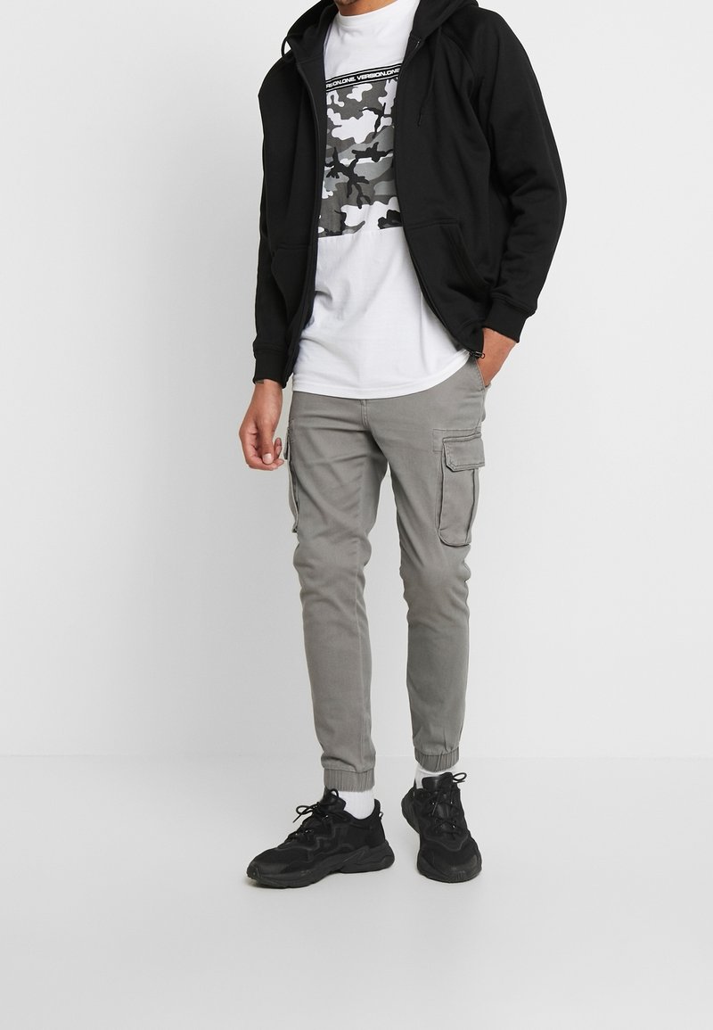 Mężczyzna w czarnej bluzie z zamkiem, białej koszulce w kamuflażu, szarych joggerach cargo, białych skarpetkach i czarnych sneakersach stojący na białym tle.