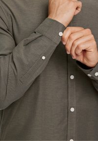 Olijfgroene blouse met lange mouwen, voorzien van een knoopkraag, witte knopen en een gladde stofstructuur. Zichtbare knoopmanchetten en placketdetails.