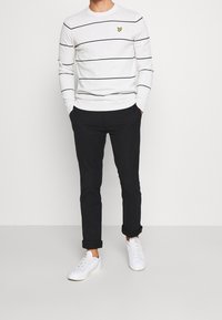 Maglione grigio chiaro a righe con strisce nere, con un piccolo emblema giallo. Indossato con pantaloni neri e sneakers bianche.