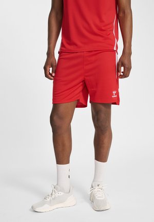 Mann trägt rote Sportshorts und passendes rotes Oberteil, steht mit weißen Sportschuhen und weißen Socken vor einem schlichten hellen Hintergrund.