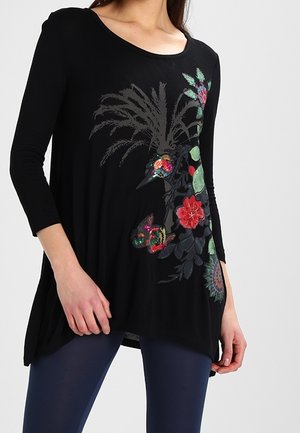 Žena nosící černou tuniku s dlouhým rukávem, zdobenou barevnou květinovou a ptáčkovou výšivkou, v kombinaci s tmavě modrými legínami, stojící před jednoduchým pozadím.