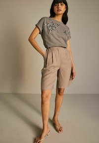 T-shirt gris à manches courtes avec trois appliqués en forme d'étoile, associé à un short beige plissé et des sandales en toile tan. Tissu lisse, coupe décontractée.