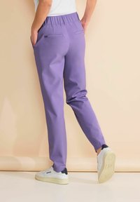 Persoon draagt paarse broek en witte sneakers, staat met handen in zakken tegen een beige achtergrond.