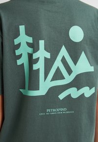Groene katoenen t-shirt met lichtteal geometrische bergen, bomen en zon graphics op de achterkant. Tekst luidt "PETROLIND SINCE '89."