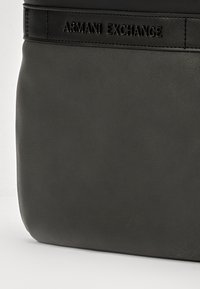 Borsa a tracolla in pelle grigia con una superficie liscia e una leggera lucentezza. Presenta il marchio "Armani Exchange" in rilievo su una tracolla nera.
