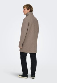 Manteau en laine beige à col montant, coupe ajustée et fentes latérales, associé à un jean foncé et des baskets noires.