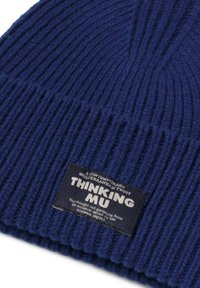 Gorro de punto acanalado en azul marino con una etiqueta rectangular negra que dice "THINKING MU" y texto adicional pequeño.