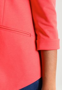 Close-up van de arm van een persoon die een koraalkleurig colbert met opgerolde mouwen en een blauwe spijkerbroek draagt, tegen een neutrale achtergrond.