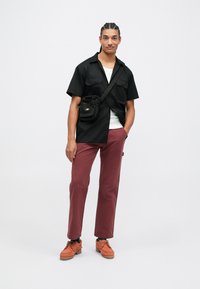 Jovem com cabelo trançado, a usar uma camisa preta de manga curta, uma t-shirt branca por baixo, calças castanhas, sapatos laranja e uma bolsa preta atravessada.