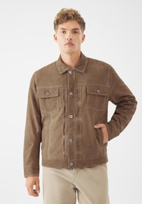 Hnědá semišová bunda s knoflíkovým zapínáním, dvěma náprsními kapsami a bočními kapsami. Má kontrastní bílé švy a klasický design límce.