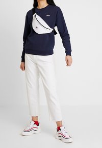 Tmavě modrá mikina s bílým logem FILA, bílá ledvinka s černým zipem, bílé zkrácené džíny a robustní bílé tenisky s červenými prvky.