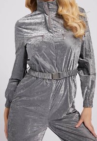 Mono gris metálico con un alto cuello, bolsillos en el pecho con cremallera, cintura elástica y tejido texturizado. Cuenta con mangas largas y puños.