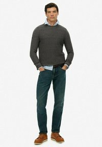 Szary sweter z dzianiny nałożony na jasnoniebieską koszulę z kołnierzykiem, w zestawieniu z ciemnymi dżinsami i brązowymi butami sznurowanymi. Prosty design z luźnym krojem.