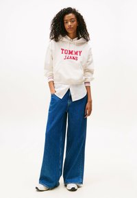Hoodie crema con "TOMMY JEANS" in rosso, sovrapposto a una maglietta bianca. Abbinato a jeans blu a gamba larga e sneakers bianche.