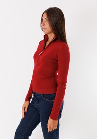 Maglione rosso con zip, dal design aderente, texture a costine e maniche lunghe. Presenta una zip parziale e motivi a costine variati sui lati.