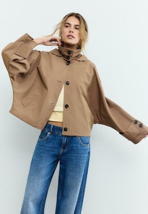 Kobieta poprawiająca kołnierz oversize'owej beżowej kurtki z dużymi czarnymi guzikami, ubrana w jasną bluzkę i niebieskie dżinsy na jednolitym tle.