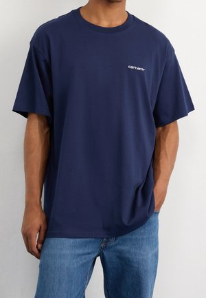 Mężczyzna w granatowym t-shircie Carhartt z krótkimi rękawami i jasnoniebieskich dżinsach, stoi z jedną ręką w kieszeni.