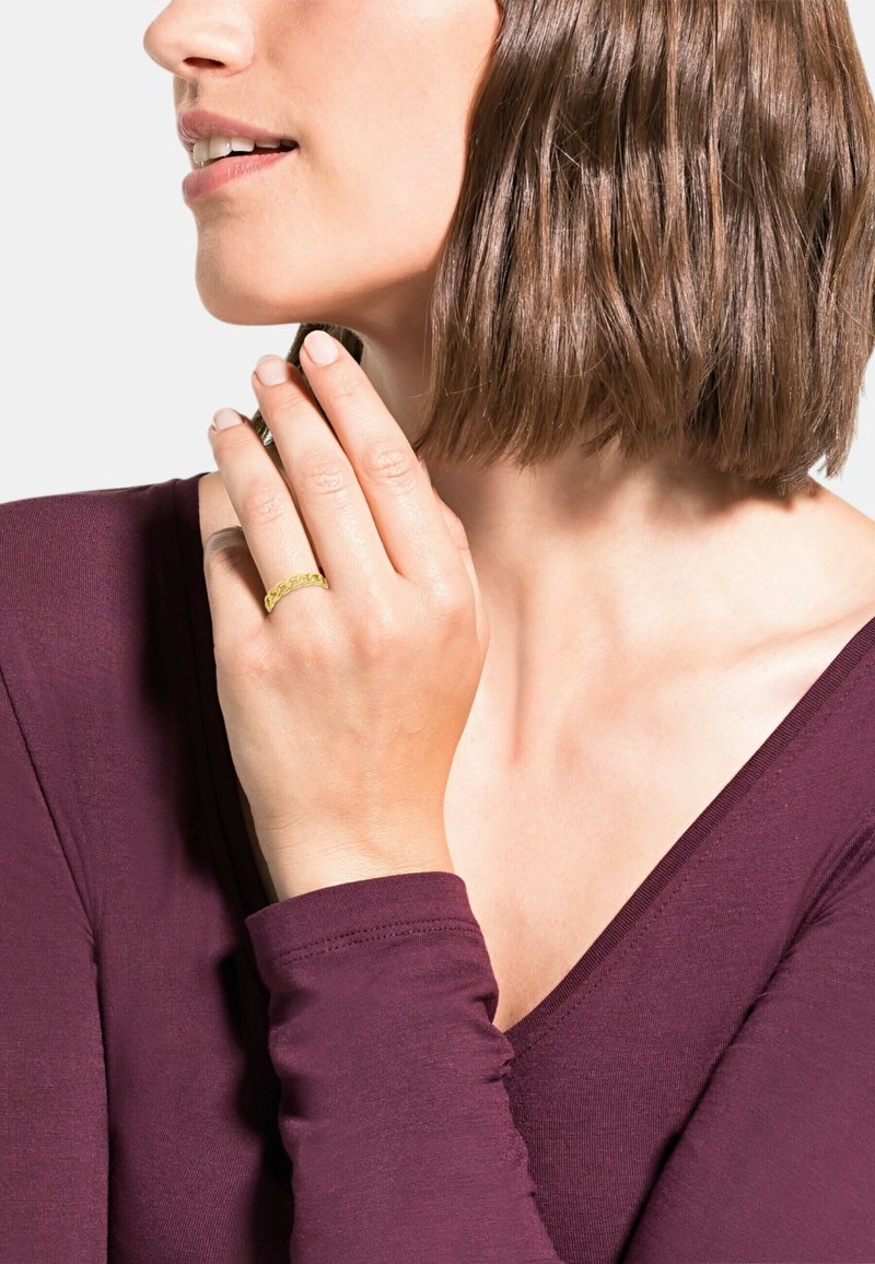 Anello in oro su una mano con pelle chiara, indossando un top a maniche lunghe di un profondo color porpora. I capelli sono castani e di lunghezza spalla.