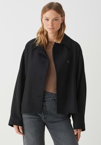 Cappotto in misto lana nero con collare, design doppiopetto e grandi bottoni. Presenta una vestibilità ampia e una texture morbida. Indossato sopra un top marrone.