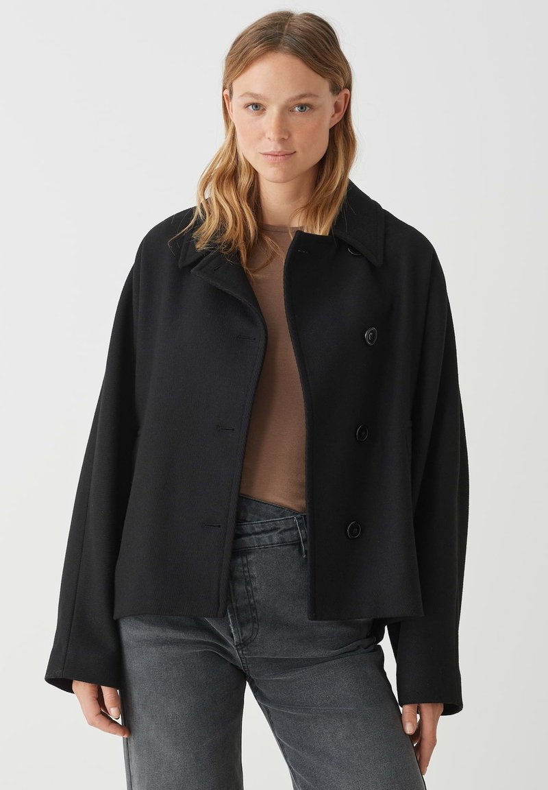 Cappotto in misto lana nero con collare, design doppiopetto e grandi bottoni. Presenta una vestibilità ampia e una texture morbida. Indossato sopra un top marrone.