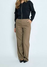 Donna con maglione nero a maniche lunghe e zip, pantaloni beige a gamba larga, scarpe nere a punta, in piedi con una mano in tasca.