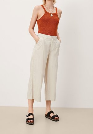 Femme portant un haut en tricot sans manches couleur rouille, un pantalon beige large et court, des sandales noires à boucle, et des colliers dorés superposés, mains dans les poches.