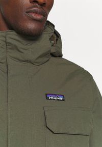 Olive green hooded jacket made of waterproof material, featuring a front pocket and a Patagonia logo patch on the chest.
