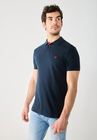 Polo shirt bleu marine à manches courtes, avec un plastron à deux boutons et des accents rouges au niveau du col. Doté d'un petit logo sur la poitrine.