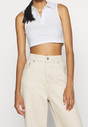 Kobieta ubrana w białą, bezrękawnikową, krótką koszulkę polo oraz beżowe spodnie z wysokim stanem, zapinane z przodu na guziki i posiadające kieszenie, stojąca na jednolitym tle.