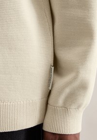 Pull léger beige avec un tricot texturé, des poignets et un ourlet côtelés. Présente une petite étiquette sur le côté avec le nom de la marque.