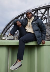 Chaqueta negra acolchada, forro polar beige, pantalones oscuros, gorro gris, zapatos de senderismo grises y naranjas, sentado en una estructura verde con un puente al fondo.