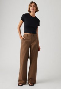 Schwarzes, kurzes T-Shirt kombiniert mit weiten, braunen Hosen. Die Hosen verfügen über Taschen und einen glatten, strukturierten Stoff. Schwarze Sandalen sind inklusive.