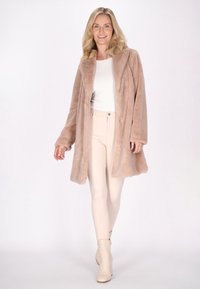 Lungo cappotto in pelliccia sintetica morbidissima di colore beige chiaro, abbinato a pantaloni attillati chiari e stivaletti alla caviglia. Presenta una linea dritta e chiusura con bottoni frontali.