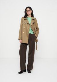 Μπεζ διπλής σειράς καμπαρντίνα με μεγάλα κουμπιά, σε συνδυασμό με πράσινο πουλόβερ και καφέ παντελόνι φαρδιάς γραμμής. Μαύρα παπούτσια ολοκληρώνουν την εμφάνιση.