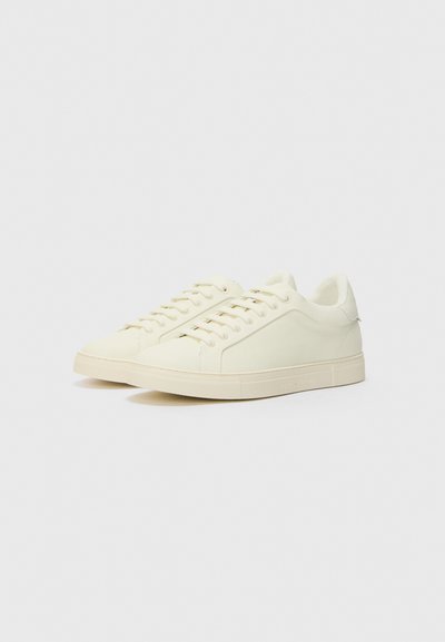 White sneakers made of smooth, soft material with a rounded toe, flat soles, and tonal laces. Side accents feature minimal branding.