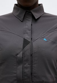 Detailný záber na tmavosivú košeľu na gombíky s ostrým golierom, čiernym sieťkovaným detailom pri gombíkoch a malým modrým trojuholníkovým logom na hrudi.