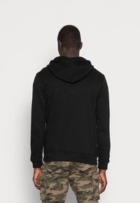 Felpa nera con cappuccio e texture morbida, dotata di tasca a canguro e polsini a costine, indossata su pantaloni cargo camouflage.